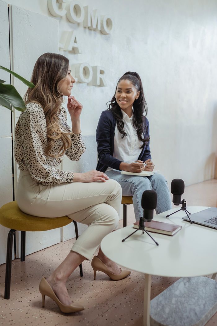 Positive multiracial female colleagues sitting on chairs near table with microphones and laptop while recording audio program in modern studio
