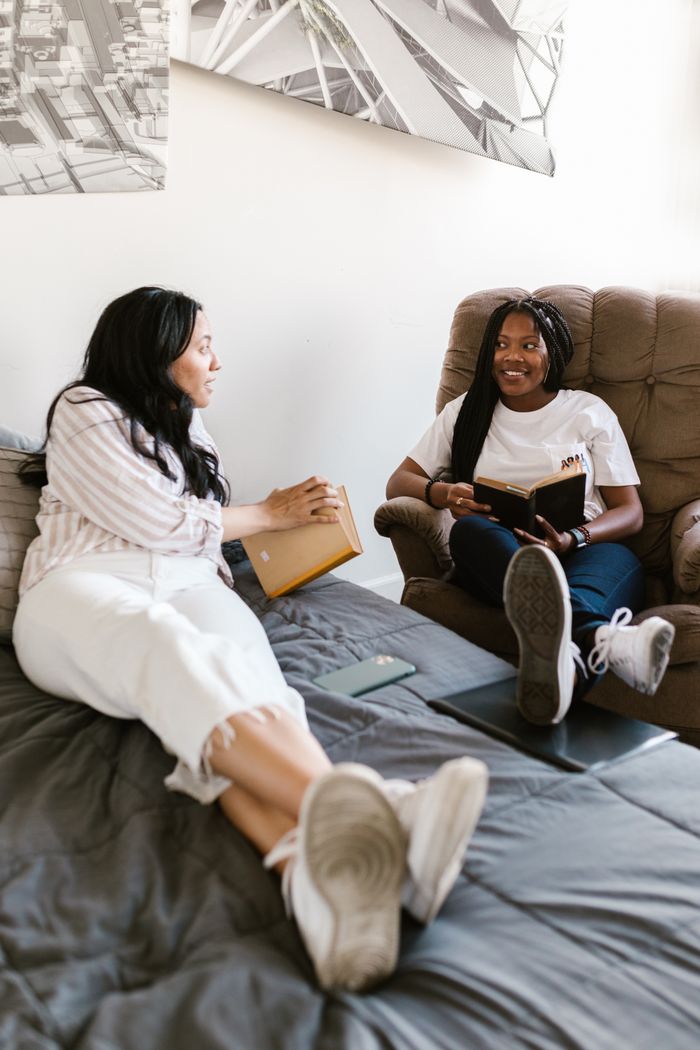 Two women studying and discussing books in a cozy indoor setting, smiling and relaxed.